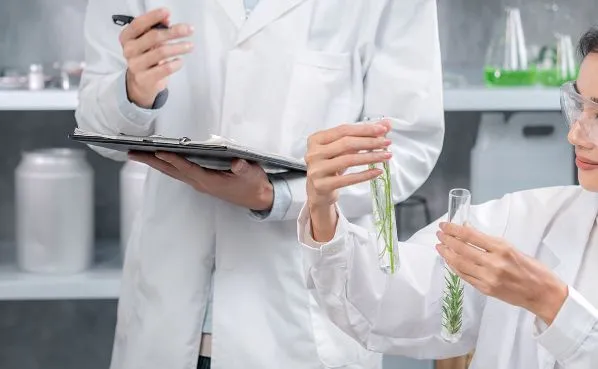 白衣を着た研究者が、植物の入った試験管を確認しながら記録している様子。